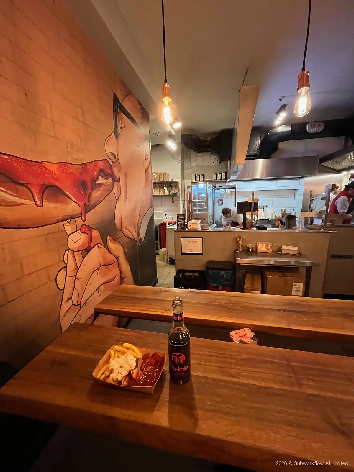 Currywurst and chips with a bottle of cola at Curry 61, under a mural of a man eating a sausage