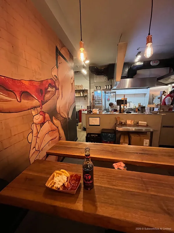 Currywurst and chips with a bottle of cola at Curry 61, under a mural of a man eating a sausage